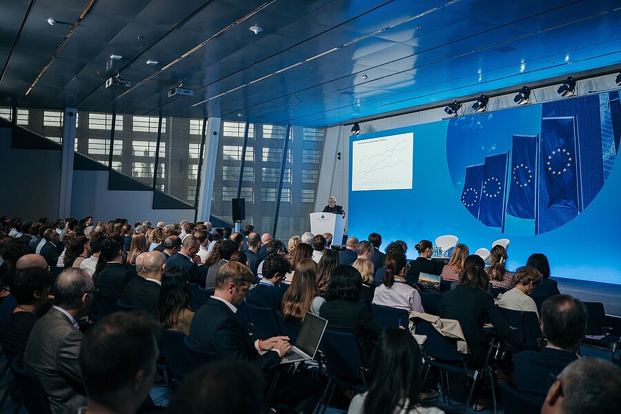 Vortrag vor vollem Saal im Konferenzzentrum der Europäischen Zentralbank, im Hintergrund Europaflaggen auf einer Leinwand